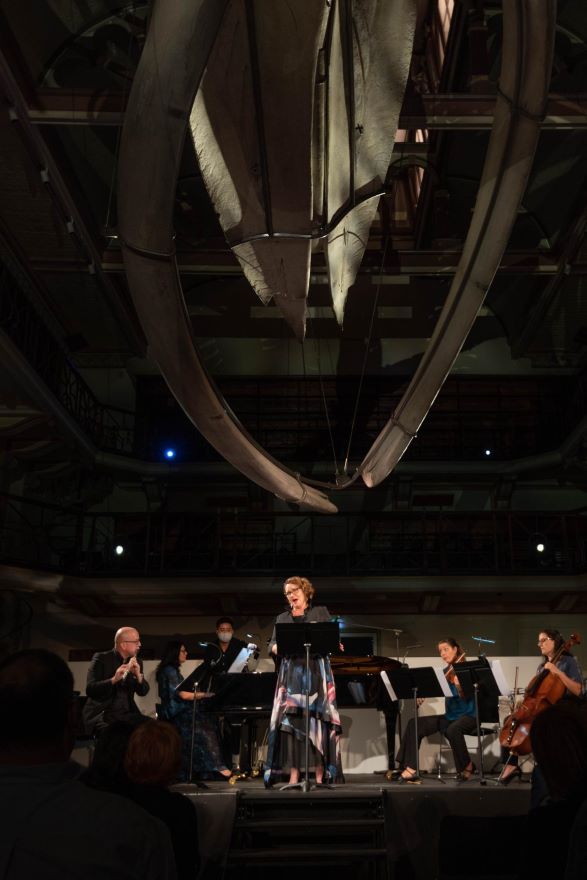 An image from the Sea Pictures performance at Hacket Hall. Pictured a large whale skeleton hangs from the ceiling of a hall, dimly lit, and on a stage below a group of 4 musicians and a singer perform