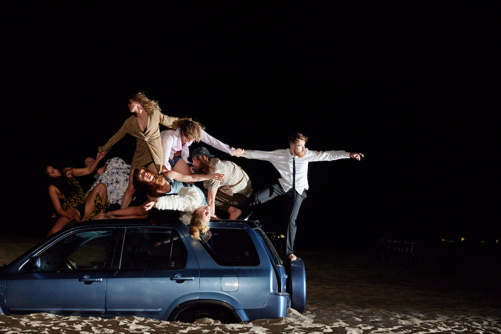 Promotional image for the performance The Ninth Wave, pictured a group of dancers are on top of a car. The car appears to be stranded in the middle of the sand.