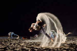Promotional image for the performance The Ninth Wave, pictured a man is dancing on a sandy beach, throwing himself forward with a bright floodlight illuminates him as he moves, throwing sand in an arc above him