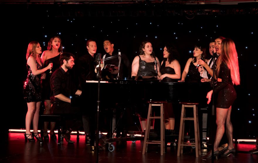 The cast of Midnite Youth Theatre Company's show The World Goes 'Round. Pictured a group of black-clad singers gather around a grand piano to sing