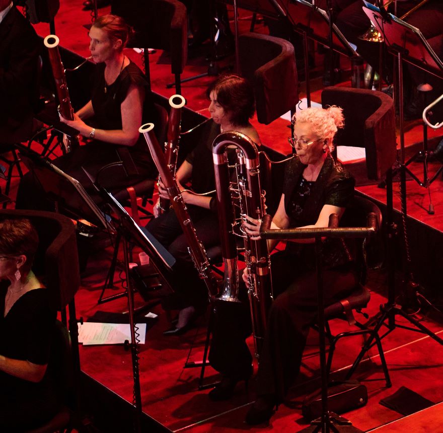 WASO contrabassoonist Chloe Turner sits on a stage playing surrounded by other orchestral musicians. The stage is lit with a wash of red light
