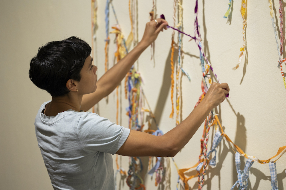 We hold you close exhibit artist Katie West, she stands and works on an art piece made of coloured string.
