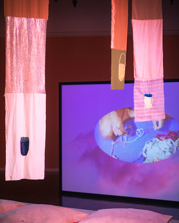 An exhibit from the We hold you close exhibition, pictured is fabric banners hang in front of a screen that is filled with a pink cloud and purple sky. In the centre, an oval features a person making string. We can't see their face, just their legs and hands.