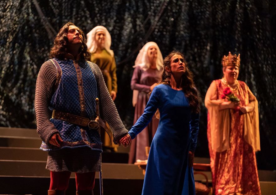 A man wearing chainmail and a sword holds hands with a woman in a long blue dress. Behind them on the stage are other characters in medieval costume