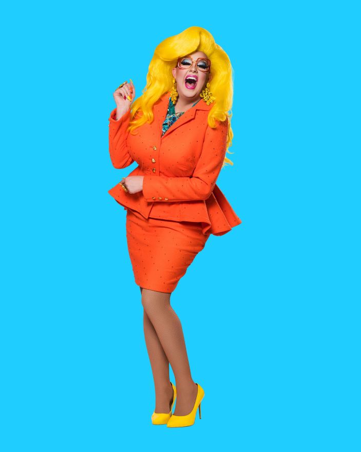 A person dressed in a yellow wig and orange business suit poses in front of a blue screen