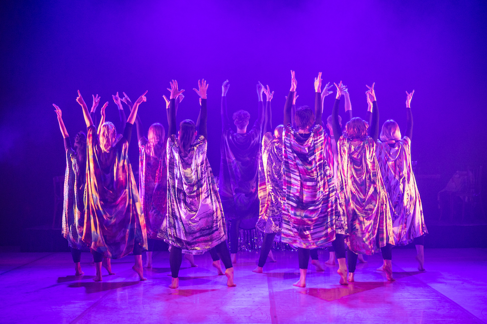 A group of dancers in colourful robes. They stand with their backs to the camera, the arms extended to the sky, seemingly joyfully.