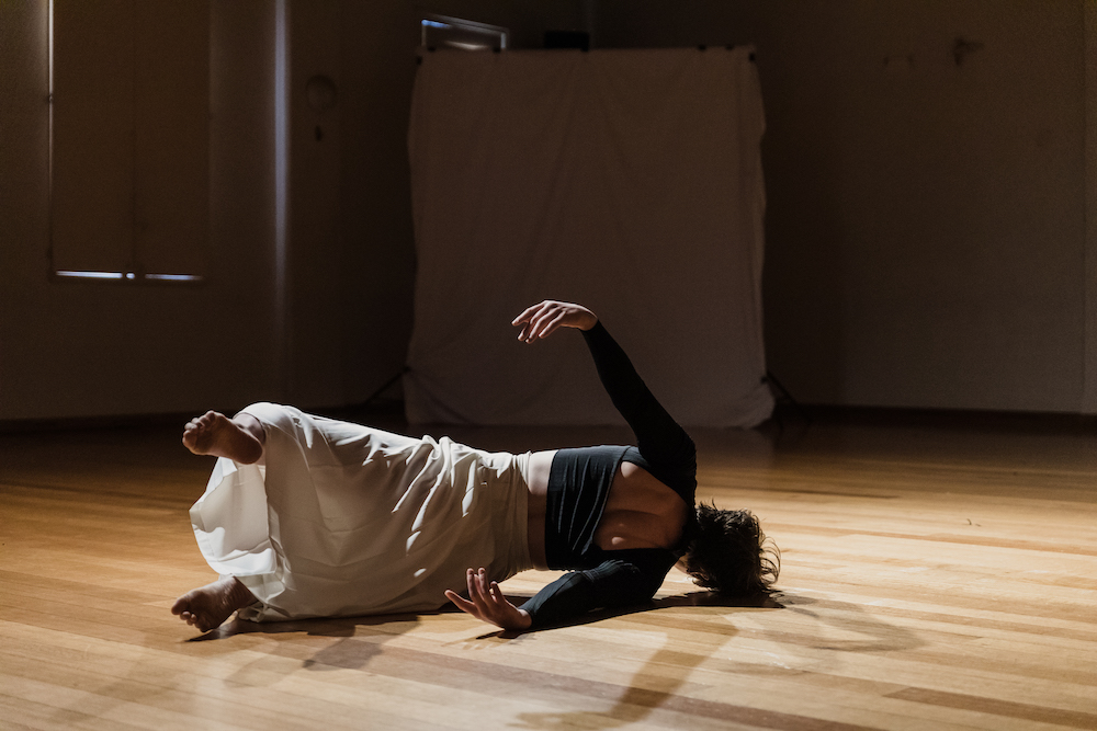 Sarah Chaffey performing in "Short Cuts". She lies on her side, facing away from us, her torso held off the ground and her arms and legs extended behind her. She wears a black fitted top with a window cut out to show her shoulder blades and cream culottes.