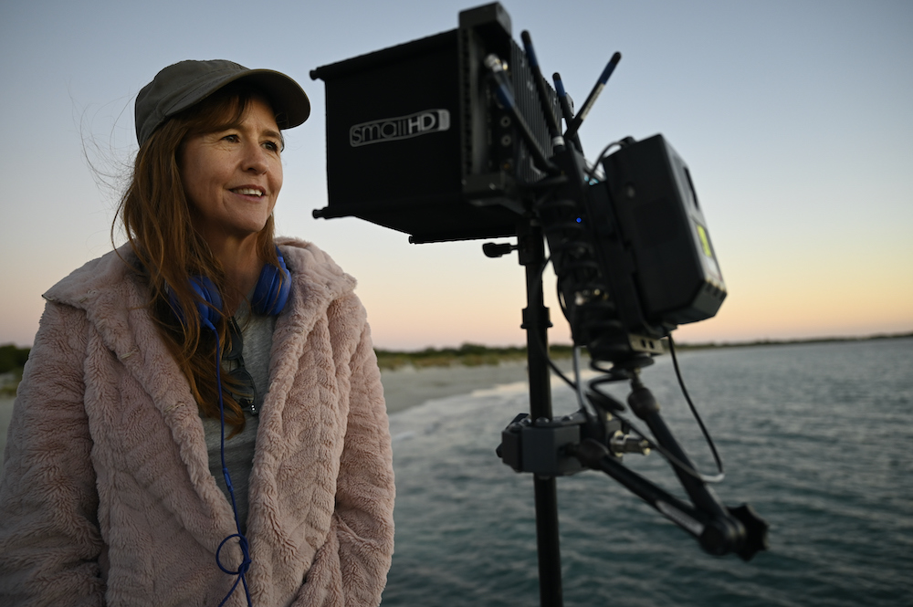 Writer and director of 'How to Please a Woman' Renee Webster stands next to some film equipment, on the beach. She is wearing a big furry coat and a cap and is gazing into the distance with a smile on her face.