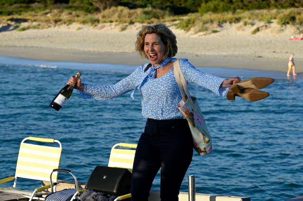 Sally Phillips is on the set of 'How to Please a Woman. She stands on what looks like a jetty, holding her shoes out in one hand and a bottle of champagne in the other. She looks like she's excited to celebrate something. 