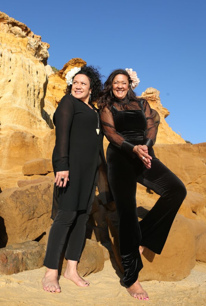 Two women with olive skin and long dark hair stand on yellow sand with yellow limestone rocks and a bright blue sky behind them. They are wearing black formal clothes and smiling