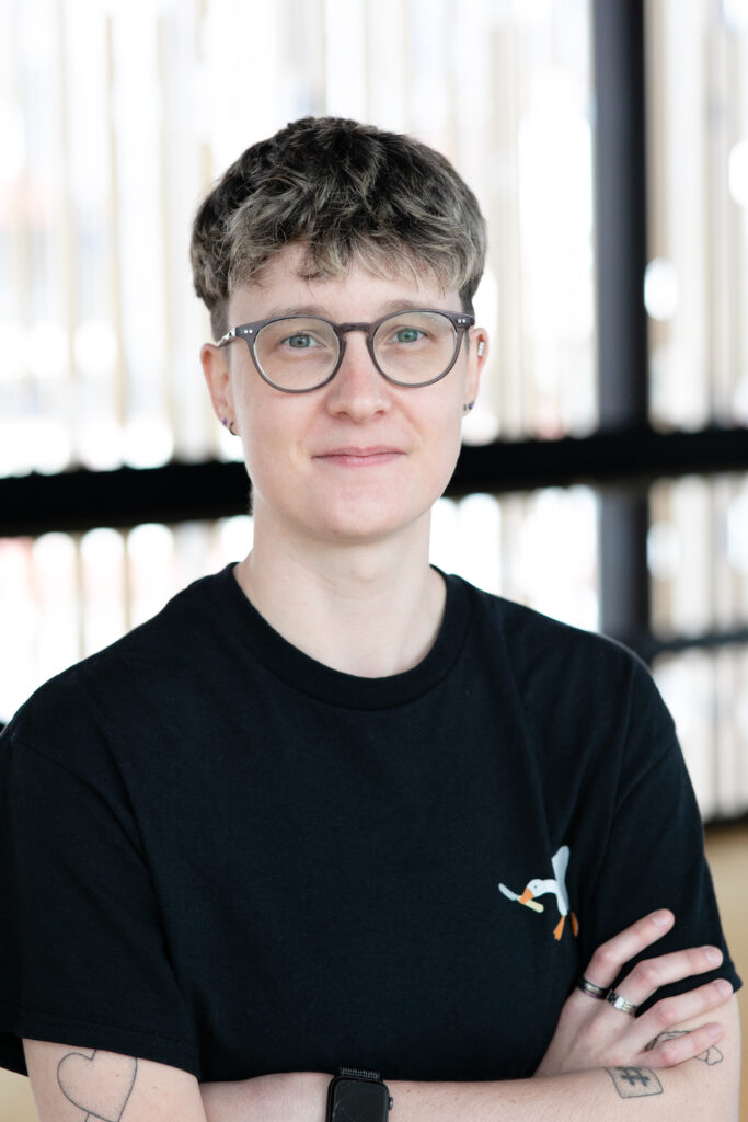 A portrait of Liz Newell. They have short hair, and they're wearing glasses and a black t-shirt with a small picture of a goose holding a knife in its beak. Their arms are folded.