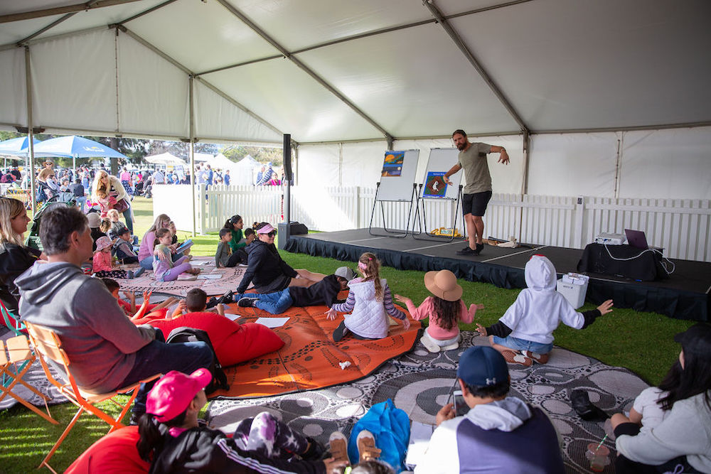 Love to Read Local. Groups of families with young children are inside a big marquee sitting on blankets and low chairs as they watch a man on a low stage. He has his legs crossed and his arms outstretched as if he is acting out a story