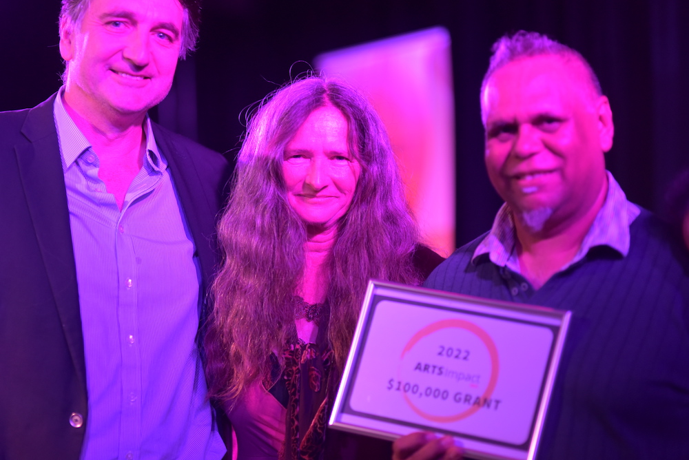 James Boyd, Vivienne Robertson and Kado Muir at the Arts Impact WA event. They are bathed in a pink light and the man holds a certificate.