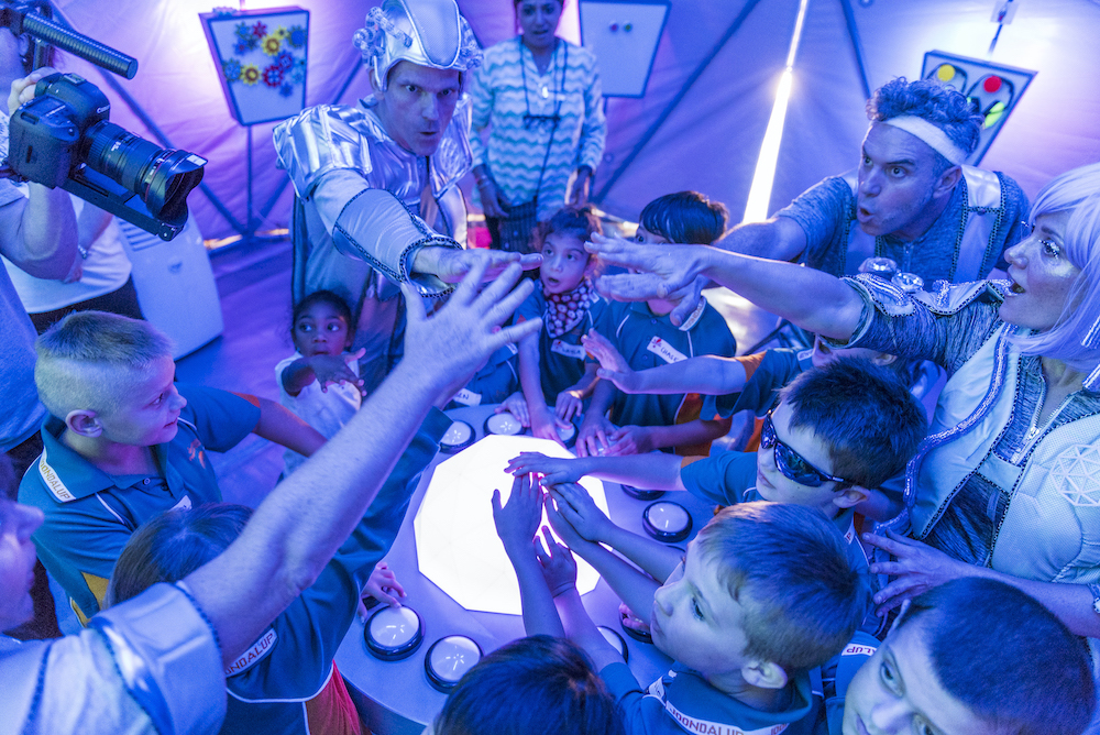 A group of children and adults clustered inside what looks like a spaceship. Everyone's hands are stretched over a brightly lit hub.