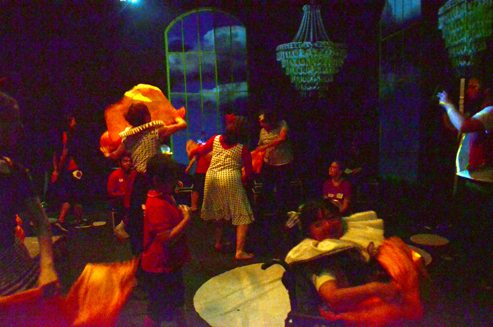Children and adults swirl around a room in Wonderbox. There are unlit chanderliers and cloud-filled windows. The adults wear giant ruffs.
