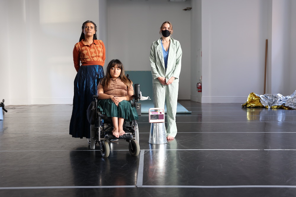 Three performers are in a dance studio. Two stand and one sits in a wheelchair. One wears a long blue shiny skirt and orange patterned sweater, one wears a pale coloured suit and one wears a brown t-shirt and green skirt. In the background we can see a sheet of lino and a crumpled length of some kind of foil like material.