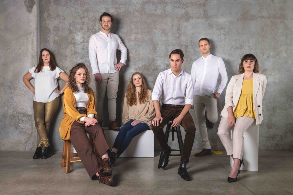 A photo of the Giovanni Consort for the July Gig Guide outlining what's on in Perth. Seven singers pose for a photo, standing or sitting casually. They wear predominantly neutral colours with the odd splash of yellow.
