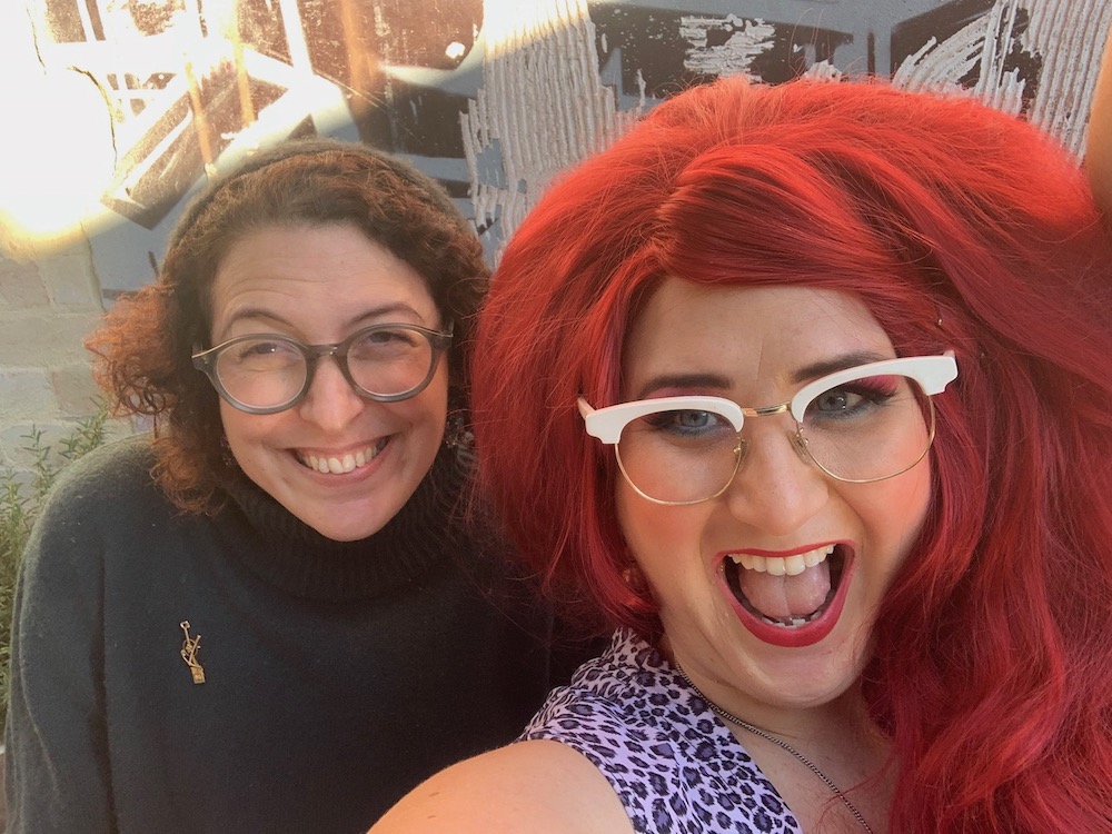 Two women grinning for a selfie. One looks very excited, the other looks even more excited. One looks fairly ordinary, the other has bright red bouffy hair and a lot of 80s style makeup.