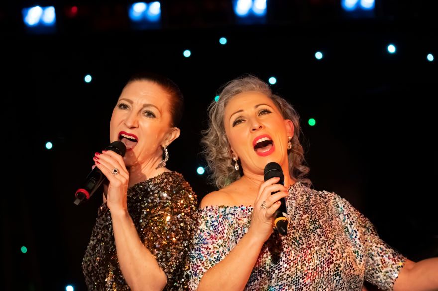 Two women in sequined dresses and holding microphones sing shoulder to shoulder under bright spotlights.