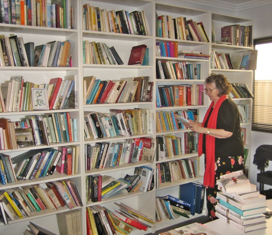A woman wearing black with a red scarf stands holding a book alongside a floor to ceiling bookshelf which is filled with books