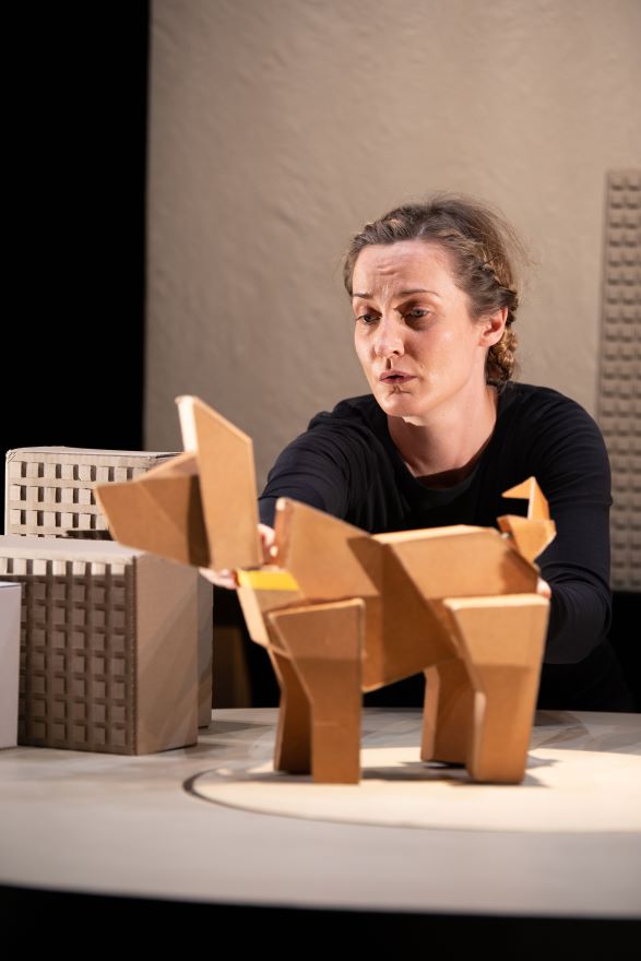 A cardboard dog stands on a table. Behind him a woman watches with a pensive face.