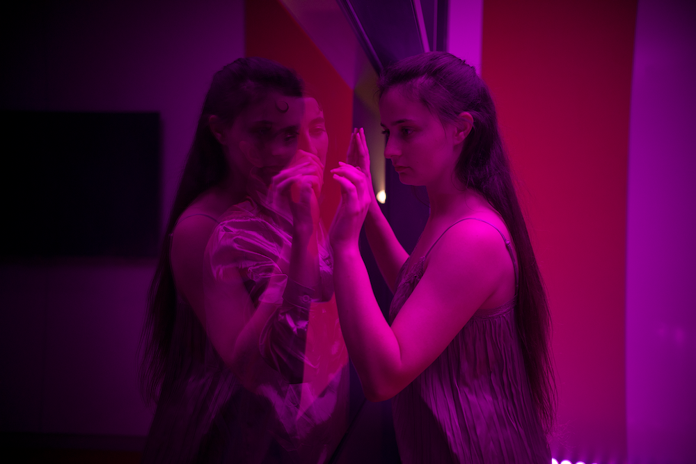 A young woman peers through a glass window and a person on the other side who is obscured by her reflection. The scene is bathed in magenta light.
