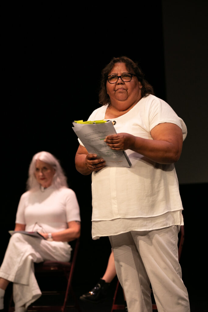 A First Nations woman dressed in white stands, holding a script. In the background, a white woman, also dressed in white, sits on a chair, a script resting on her legs.