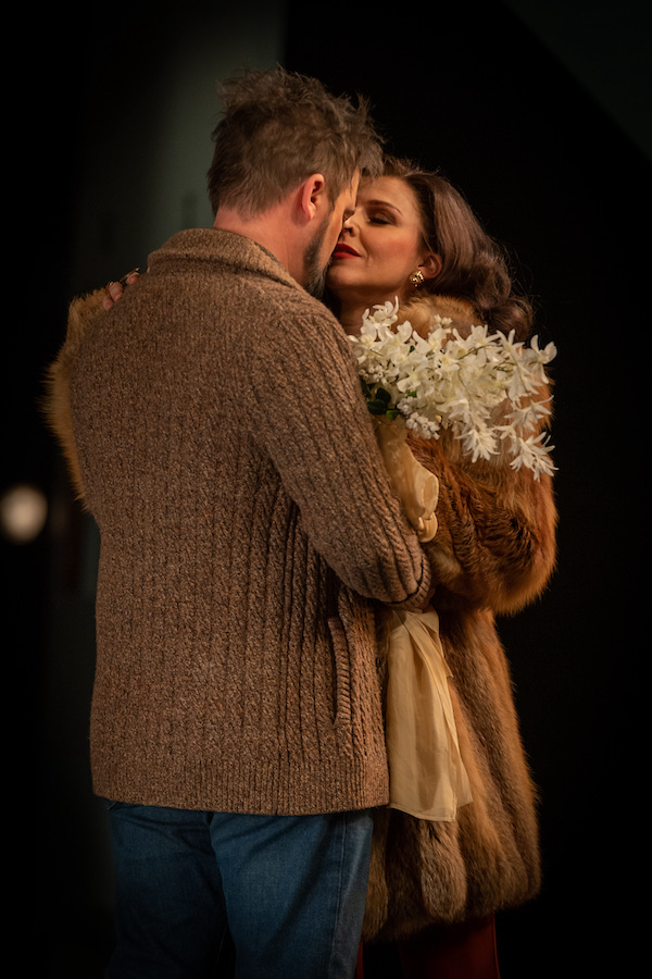 Tosca. A man in a knitted brown cardigan and a woman in a brown coat holding flowers are locked in a passionate embrace