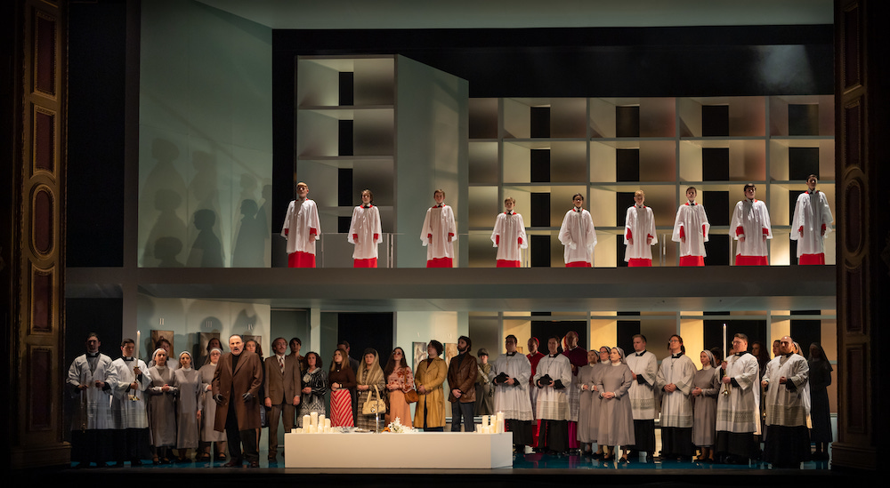 Tosca. We see a two-storey set on a stage. The top level has members of a choir in red and white robes. below them are a large group of performances, some in black and white robes   