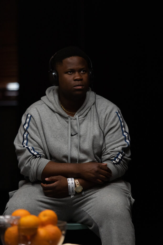 Tinashe Mangwana as Christopher in Blue/Orange sit with a slightly belligerent expression, his arms loosely crossed, headphones covering his ears.