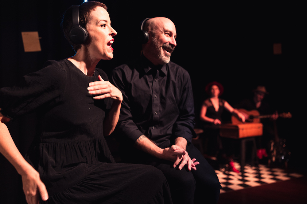 Two performers wear headphones and smile at an unseen audience. It looks like they're talking or singing.