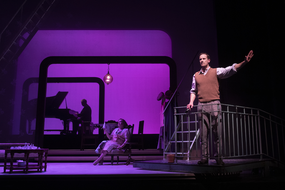 A man stands near in a gate at the foreground, raising his arm; in the background, a woman in a chair and a man at a grand piano are bathed in soft purple light.