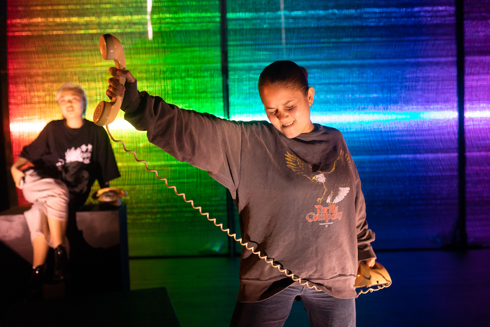 Cream of the crop:A young woman in a sweatshirt holds a landline telephone receiver in one hand, like it is a trophy or a prize, and the telephone itself in the other. The background is coloured in a rainbow spectrum.