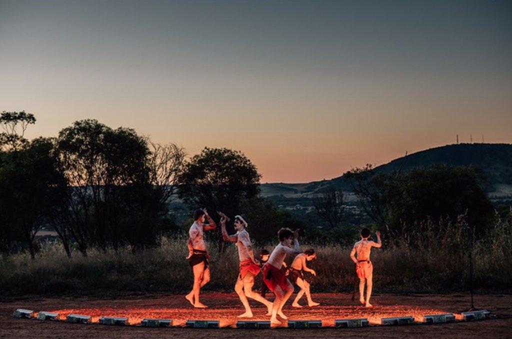 A bush setting at twilight with FIrst Nations dancers in traditional dress and paint lit by footlights