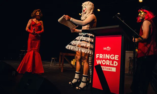Fossil fuel divestment - artists protest Fringe World's fossil fuel sponsors. Three artists dressed in elaborate red and white costumes stand on a stage next to a lectern bearing the Fringe World logo.