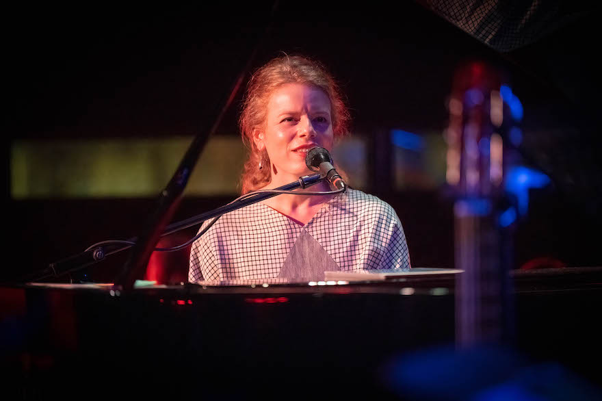 A woman in a checked top, hair pulled back, sits behind a piano leaning into the microphone. Georgie Aué is launching her new album Desert Sky.