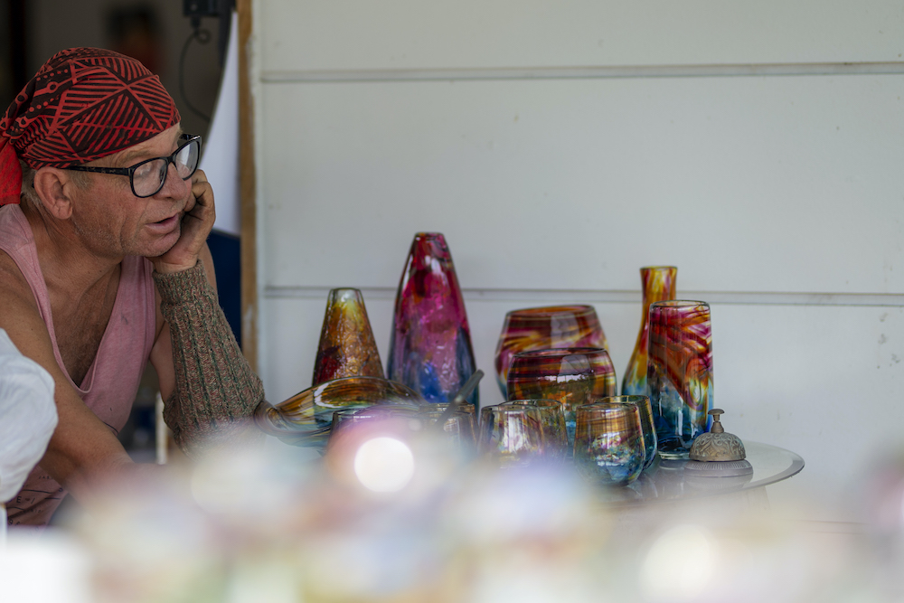 Gerry Reilly sits at a table, one arm resting next to an assortment of rainbow hued vessels.