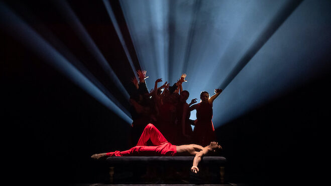 A shirtless man in red pants is lying down trying to sleep, dancers behind against shafts of light amid the darkness reflecting his tormented mind.
