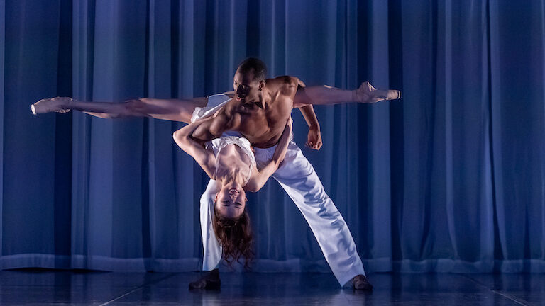 A dancer holds the White Muse in one arm, her back arched and head tipped forward, legs spread out behind him.
