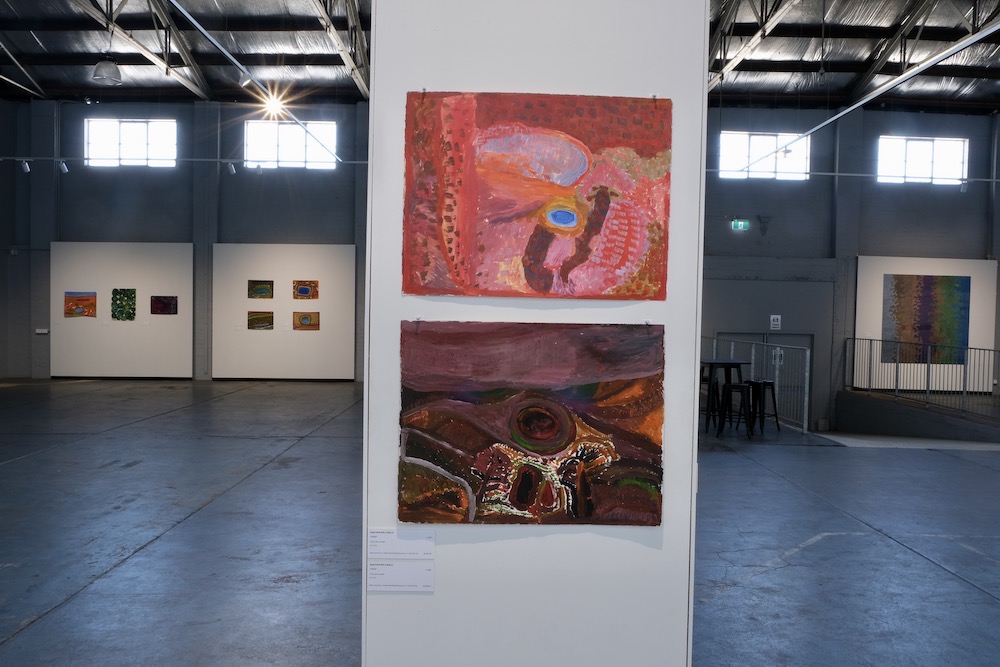 Two abstract landscape paintings on paper at 'Mangkaja 30 Years Strong', one in bright reds and pinks, the other in darker burgundies and purply-mauves.