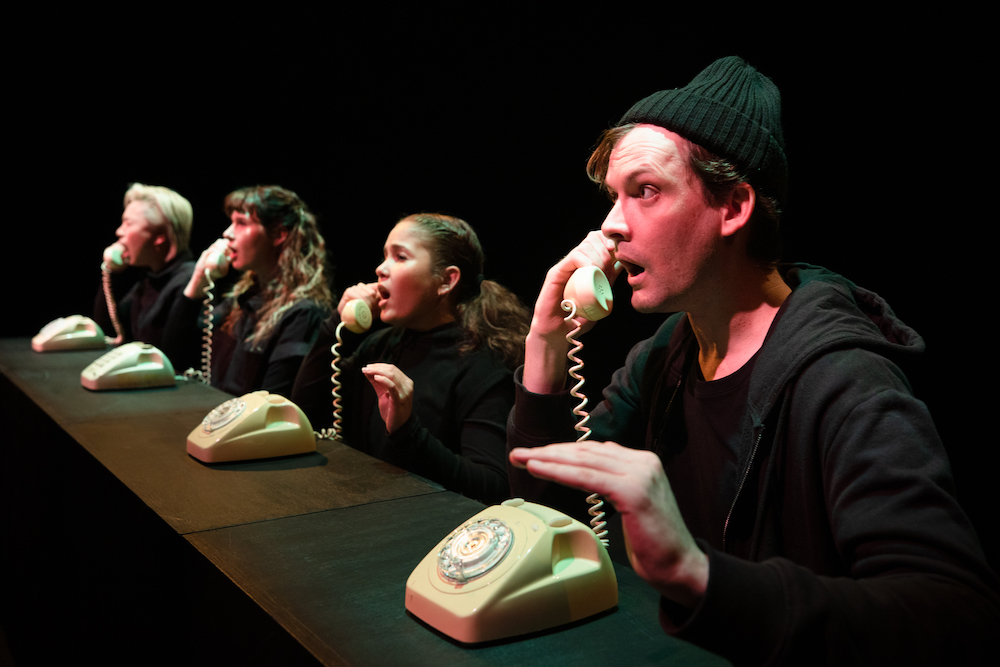 A line of four actors, clothed in black, talking on rotary dial phones.