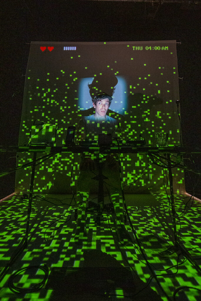 A stage is covered in squares of green light, referencing the green cursor of vintage compuers. In the centre of a screen we can see the face of a young man.