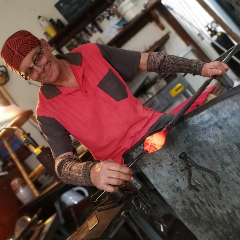 Gerry Reilly holds a metal rod - at one end is a piece of molten glass, which he is manipulating with another metal tool. He is smiling as he works.