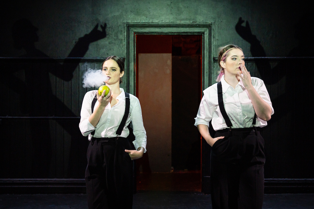 Two young women are dressed like Charlie Chaplin, in white shirts, black suspenders and black pants. One mimes smoking a cigarette, the other blows out a mouthful of smoke.