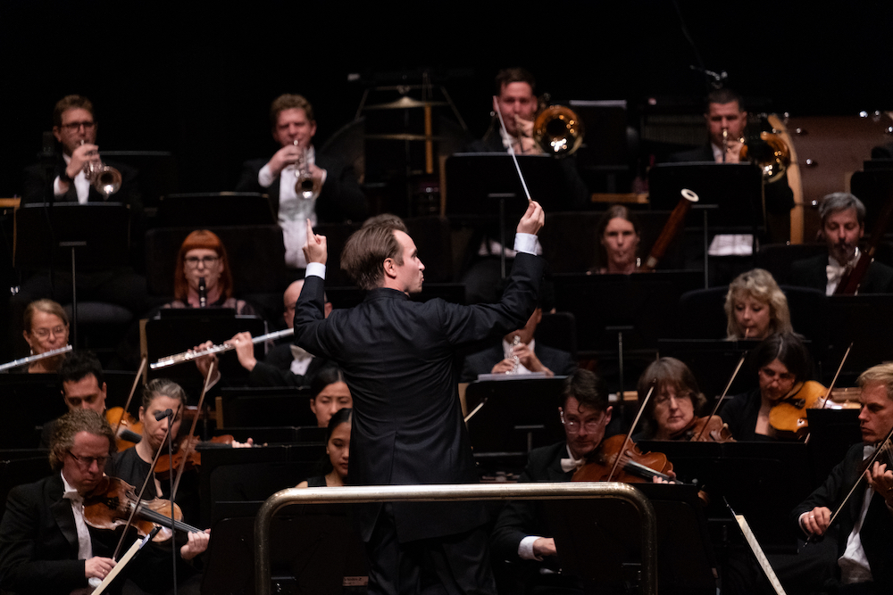 A conductor with his back to the camera raises his baton to the orchestra, musicians concentrating on their instruments. It is conductor Pietar Inkinen with the West Australian Symphony Orchestra.