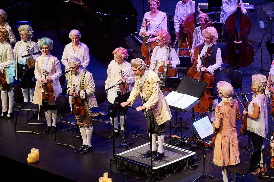 Musicians dressed in 18th century garb, including wigs, and holding various instruments, stand on the stage bowing to an audience out of sight.  They are Perth Symphony Orchestra performing Amadeus.