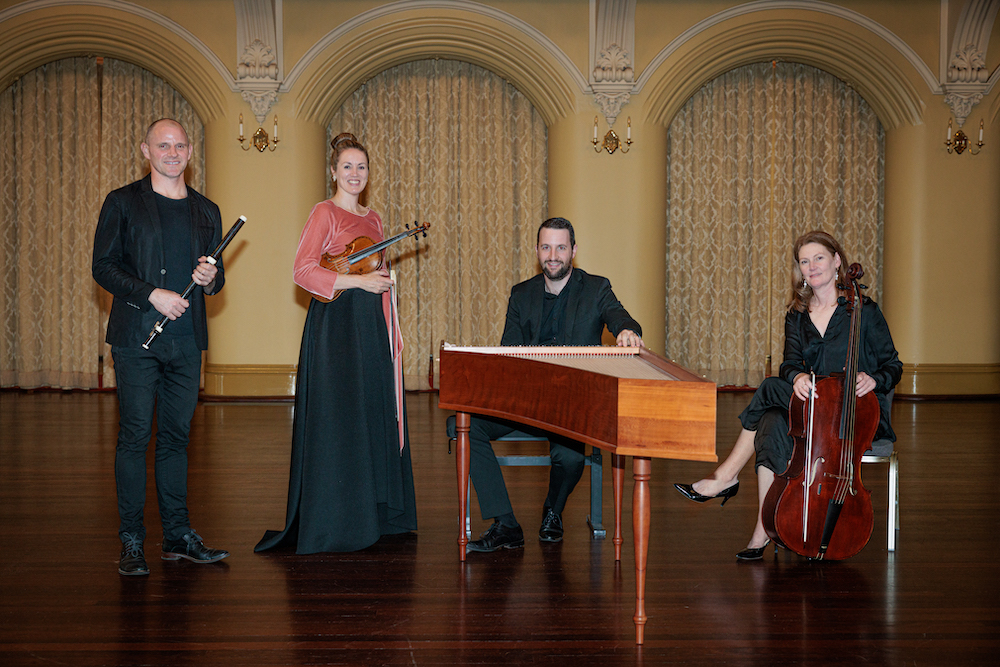 Four musicians smile at the camera, arched columns behind them. One is holding a flute, the other a violin, while the third musician is seated behind a harpsichord and the fourth,  behind her cello. They are Australian Baroque performers Andy Skinner, Helen Kruger, James Huntingford and Noeleen Wright.