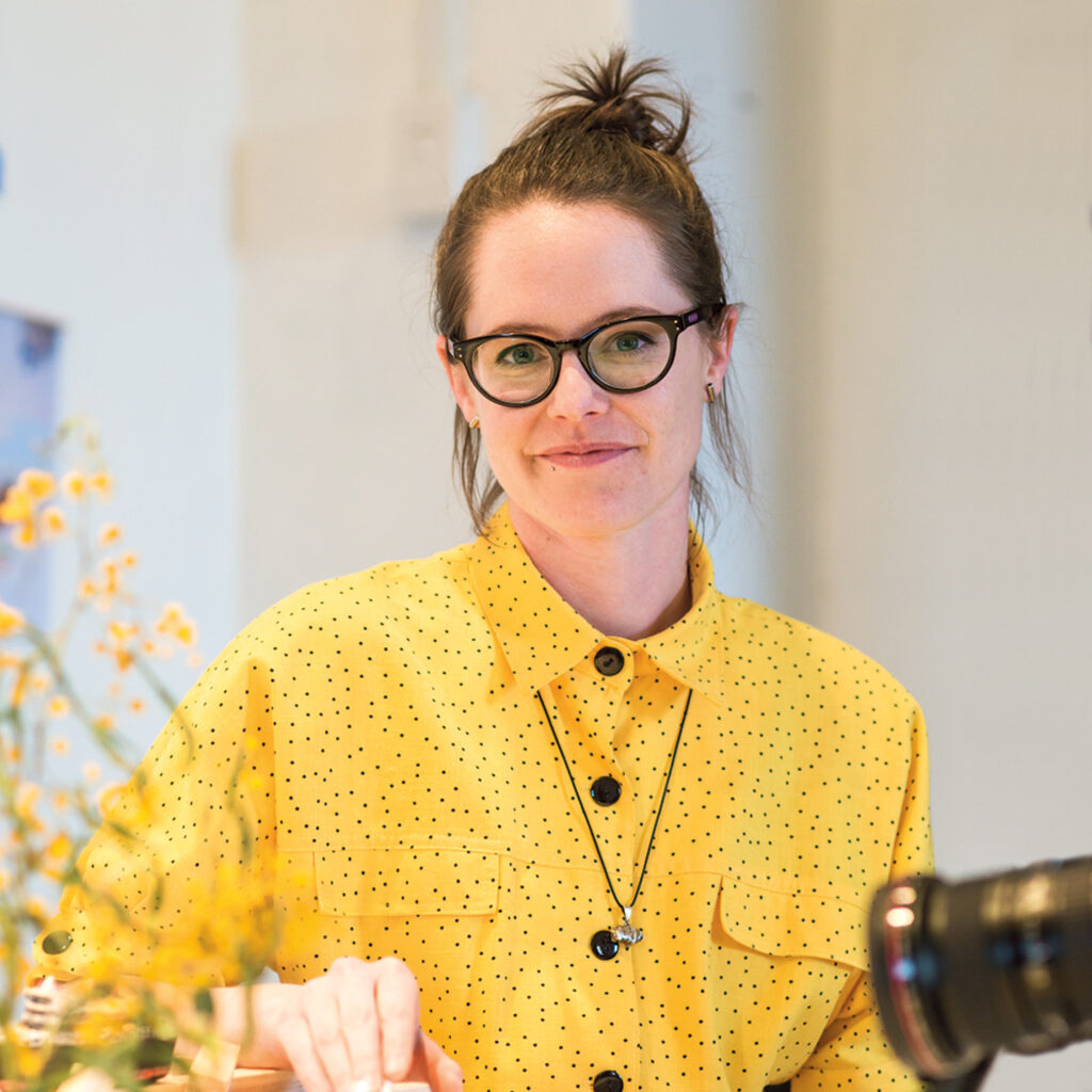 Melville Storylines promotional image: Female WA artist Amy Perejuan-Capone 
will be joining the Melville Storylines program. She smiles at the camera in a bright yellow button-up, and in the foreground is a sprig of yellow flowers.