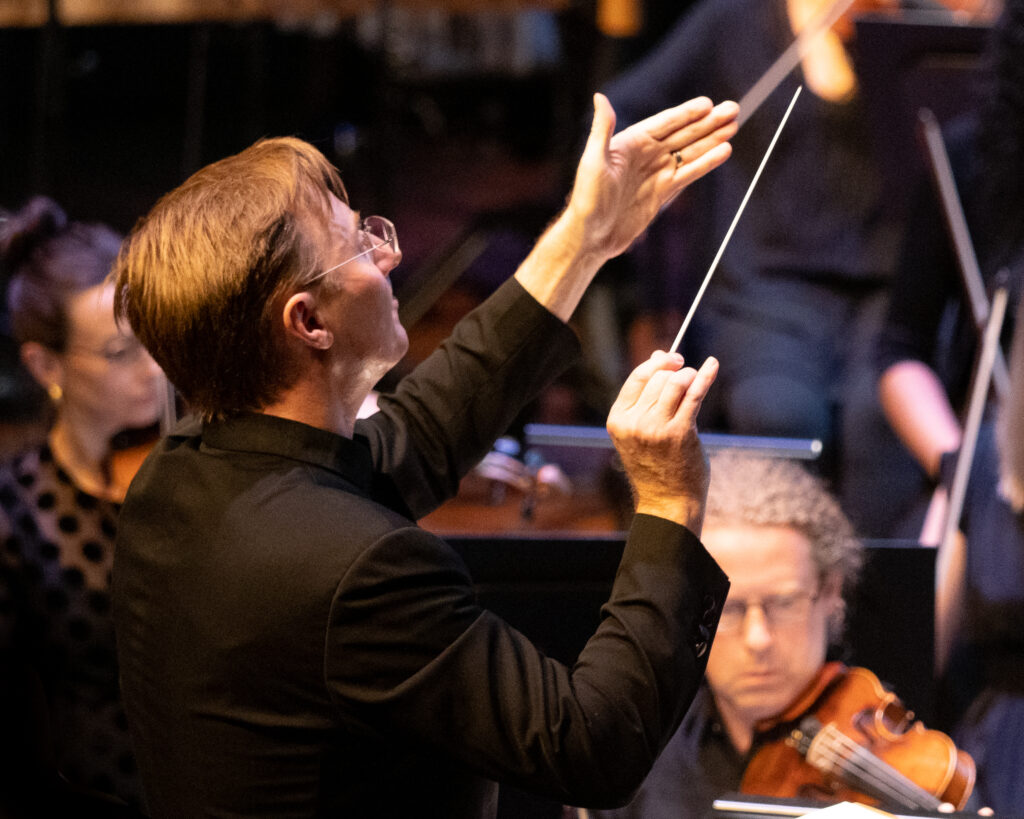 A conductor with his back to camera raises his baton to the musicians facing him. It is Benjamin Northey conducting WASO's performance of Eumeralla.