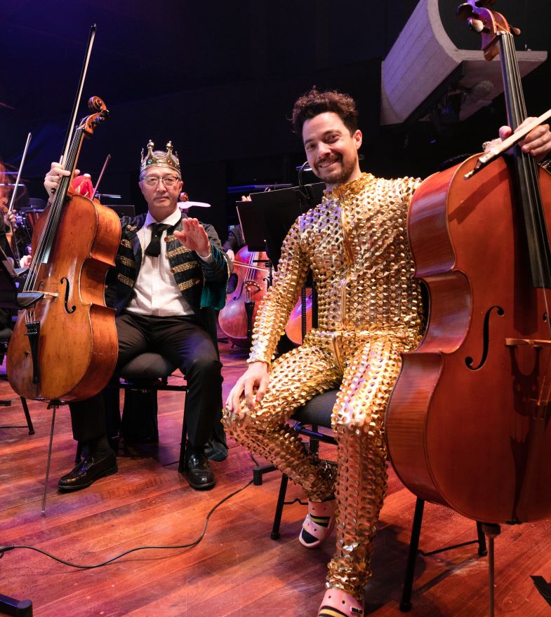 Two men in glittering costumes sit on a stage holding cellos and smiling